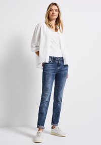 Camisa blanca ligera de botones sobre una camiseta de tirantes blanca, combinada con unos vaqueros azules desgastados que presentan detalles rasgados. Zapatillas blancas completan el conjunto.