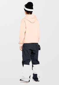 Pfirsichfarbener Hoodie mit einer Kordel am Kragen; kombiniert mit schwarzen Hosen, die im unteren Bereich weiß sind und seitliche Taschen haben; getragen mit einer schwarzen Mütze.