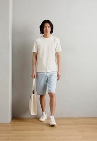 Beige Baumwoll-T-Shirt, hellblaue Jeans-Shorts, weiße Sneakers und eine beige Tragetasche. Steht auf einem Holzfußboden vor einem neutralen Hintergrund.