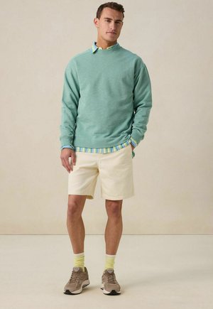 Mintgroene sweatshirt, gelaagd over een lichtblauw en gestreept overhemd, gecombineerd met crèmekleurige shorts en tan sneakers met gele sokken.
