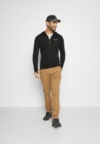 Crna zip-up dukserica s narančastim detaljima, uparena s bež cargo hlačama i crnim cipelama. Model hoda i smiješi se.