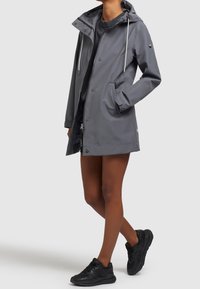 Osoba v šedém deštníku s kapucí, má přední kapsy, černé tenisky a tmavé tričko, stojí s jednou rukou v kapse.