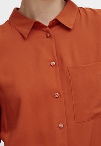 Chemise orange à manches courtes avec boutons, fabriquée en tissu lisse, présentant un col classique, une poche plaquée et des boutons nacrés.