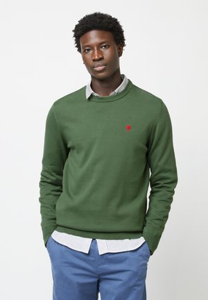 Groene gebreide trui met een ronde hals, voorzien van een klein rood boompje-logo op de borst, gedragen over een blauw-wit gestreept shirt.