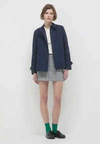 Giacca in cotone blu navy con taglio dritto, due bottoni e polsini arrotolati, abbinata a una gonna scozzese grigia e calze verdi.