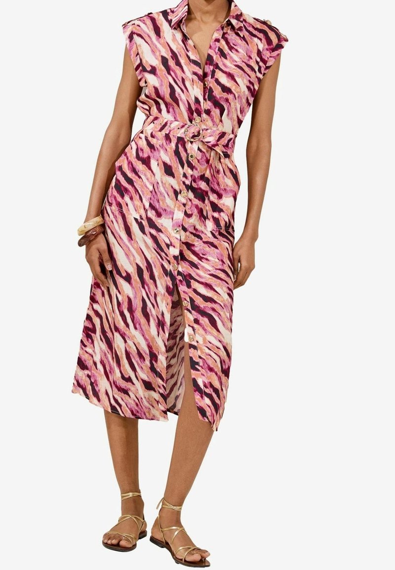 Robe midi sans manches ceinturée avec un motif abstrait rose, violet et crème, boutons sur le devant, portée avec des sandales dorées à brides et des bracelets.