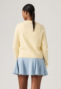 Γυναίκα με πλεγμένα μαλλιά που φοράει ένα κρεμ πουλόβερ με πλέξη καλώδιο και μια ανοιχτό μπλε πλισέ τζιν φούστα, απεικονίζεται από πίσω με φόντο ένα μονόχρωμο τοπίο.