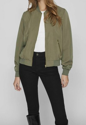 Femme portant une veste bomber vert olive sur une chemise blanche, un pantalon noir taille haute et des bottes noires à mi-mollet sur un fond uni.