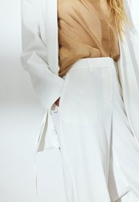 Blazer blanco ajustado con un bolsillo y mangas largas, superpuesto a una blusa beige transparente y pantalones blancos de cintura alta. Textura suave en toda la prenda.