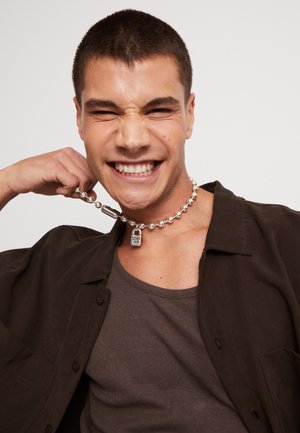 Joven sonriendo con los ojos cerrados, vistiendo una camisa oscura y una camiseta sin mangas, sosteniendo un collar de cuentas plateadas con un colgante de candado cerca de su cuello.