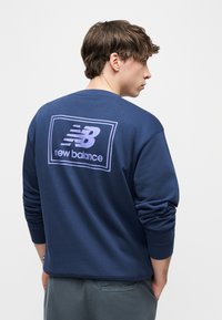 Νέος άνδρας που φοράει μπλε σκούρο φούτερ με μεγάλο ανοιχτό μοβ λογότυπο "New Balance" στην πλάτη, όρθιος με τα χέρια στις τσέπες.