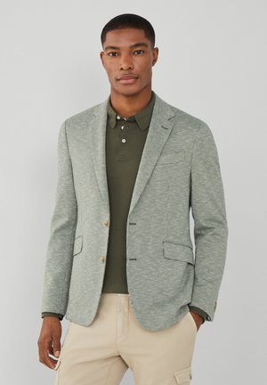 Hellgrüner Hahnentrittmuster-Blazer mit zwei Knöpfen, Reverskragen und Taschen. Kombiniert mit einem olivgrünen Poloshirt und beigen Stoffhosen. Texturiert.