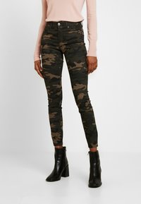 Jean skinny camouflage aux tons verts et marron, avec taille haute, cinq poches et fermeture éclair. Porté avec des bottines noires à talons.