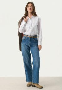 Hvid skjorte med knapper, blå high-waisted jeans, lysebrun bælte og lysebrune ankelstøvler. Modellen holder en brun jakke over skulderen.