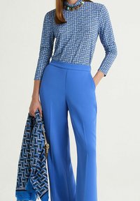 Pantalon bleu ajusté assorti à un haut bleu à manches longues à motifs. Le haut présente un design géométrique et un col montant, orné d'un col en perles colorées.