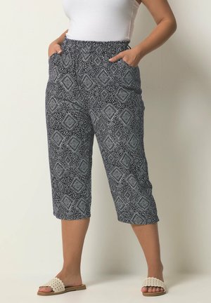 Kobieta ubrana w czarno-białe wzorzyste spodnie 7/8, biały bezrękawnik i białe klapki, stojąca z rękami w kieszeniach.