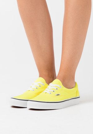 Žluté plátěné tenisky s bílými tkaničkami, kulatou špičkou a gumovou podrážkou. Obsahují malé logo Vans na boku a černý akcent na okraji podrážky.