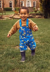 Modré nepremokavé nohavice s vzormi žltých stavebných vozidiel, nosené cez béžový pletený sveter. Čierne gumáky, pozadie z trávy.