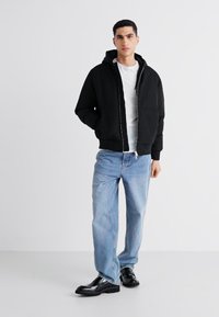 Chaqueta con capucha negra de cremallera y corte relajado, llevada sobre una camiseta blanca, combinada con jeans de color azul claro y zapatos negros. Diseño y textil sencillos.