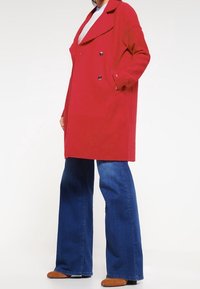 Rode dubbelzijdige jas met grote revers en twee voorzakken, gedragen over een lichtblauw top en gecombineerd met blauwe flared jeans en bruine schoenen.