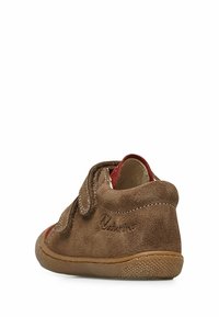 Scarpa per bambino in suede marrone con un accento di tessuto rosso e chiusura in Velcro. Forma arrotondata e suola in gomma flessibile per comfort e supporto.