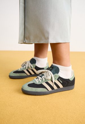 Person iført hvide sokker og Adidas Samba sneakers med sorte, grønne og cremefarvede detaljer, der står på et gulgulv.