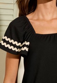 Černý top s vypouklými rukávy s krémovým zvlněným lemem, čtvercovým výstřihem a nařasením na rukávech. Měkká textura látky.