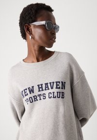 Pilkas džemperis su "NEW HAVEN SPORTS CLUB" tekstu tamsiai mėlyna spalva. Modelis dėvi skaidrius akinius nuo saulės ir sidabrinę auskarą. Minkšta medžiaga, laisvas siluetas.