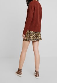 Nainen, joka on pukeutunut ruosteenväriseen neuleeseen, leopardikuvioiseen mini-hameeseen ja leopardikuvioisiin korkeisiin tennareihin, seisoo yksinkertaista harmaata taustaa vasten.