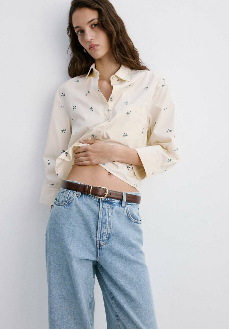 Jeune femme portant une chemise crème à fleurs boutonnée partiellement rentrée dans un jean bleu clair avec une ceinture marron, debout contre un mur blanc.
