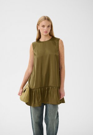 Kvinde med langt blond hår iført ærmeløs olivegrøn flæset tunika over blå jeans, holder en tekstureret beige clutchtaske mod hvid baggrund.