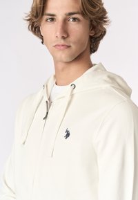 Giovane uomo con capelli castano chiaro che indossa una felpa bianca con zip e cappuccio, con un piccolo logo scuro di un giocatore di polo sul petto.