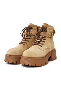 Beige suède enkellaarzen met een dikke brown zool, voorzien van veters, een zijrits en een band met een gespdetail. Gestructureerd oppervlak.