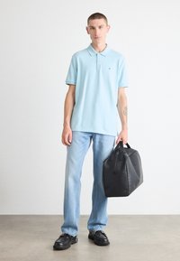 Lyseblå polo-shirt med krave, parret med lyseblå jeans og sorte lædersko. Holder en sort duffel taske med minimalistisk design.