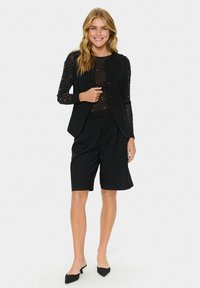 Schwarze ärmellose Weste mit drei Knöpfen, transparente schwarze Bluse mit silbernen Mustern und maßgeschneiderte schwarze Shorts, ergänzt durch spitze schwarze Schuhe.