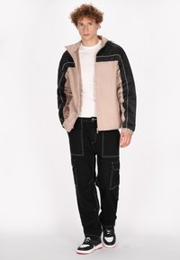 Lichte beige en zwarte jas met contrasterende stiksels, gecombineerd met zwarte cargobroeken met zakken en een relaxte pasvorm, afgemaakt met sneakers.
