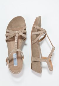 Sandales en suede beige avec des brides entrecroisées, un accent en strass au centre et un talon plat avec une fermeture à boucle.