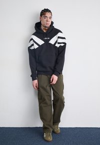 Svart hoodie med vita diagonala ränder, kenguruficka; matchad med olivgröna cargobyxor och bruna sneakers med vita ränder.