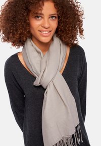 Écharpe gris clair en matériau doux, présentant une texture lisse et des extrémités à franges, portée par-dessus un pull noir à large encolure.