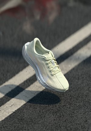 Chaussure de sport vert clair avec une tige en maille, une fermeture à lacets et une semelle amortie, montrée en l'air au-dessus d'une surface texturée sombre.