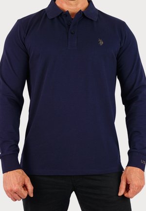 Mężczyzna w granatowej koszuli polo z długim rękawem, z małym haftowanym logo na piersi i napisem "US" na mankiecie.