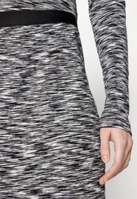 Gros plan d'une personne portant une robe tricotée à manches longues avec un motif abstrait noir et blanc et une ceinture noire.