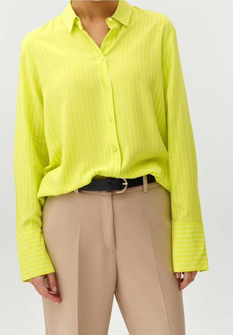 Chemise à rayures jaune vif avec boutons, rentrée dans un pantalon beige taille haute avec ceinture noire, montrée du cou aux hanches.