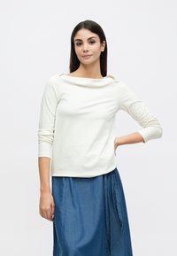 Maglia a maniche lunghe color avorio con scollo a barca e tessuto morbido, abbinata a una gonna di denim blu con dettagli plissettati.