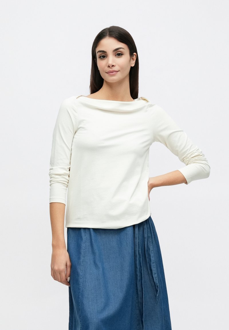 Maglia a maniche lunghe color avorio con scollo a barca e tessuto morbido, abbinata a una gonna di denim blu con dettagli plissettati.