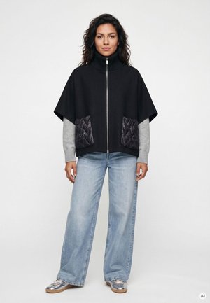 Frau trägt einen schwarzen Zip-Up-Cape mit gesteppte Taschen, graue Pulloverärmel, weite hellblaue Jeans und silberne Sneaker, steht vor weißem Hintergrund.
