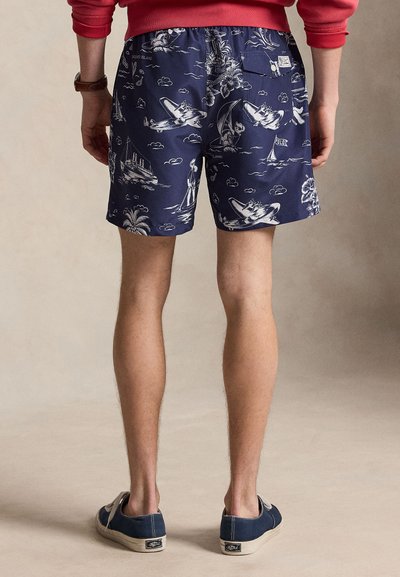 Muž v tmavě modrých šortkách s bílým retro vzorem letadel, lodí a tropických motivů, doplněných červenou mikinou a modrými teniskami.