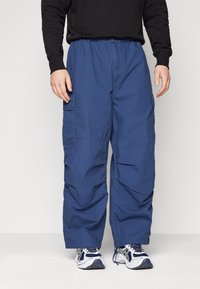 Siniset kuormahousut, jotka on valmistettu kevyestä kankaasta. Housuissa on joustava vyötärö, sivutaskut ja rento istuvuus. Yhdistetty valkoisiin urheilukenkiin.