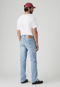 Vaqueros de mezclilla de color azul claro con un corte relajado, textura desgastada, parche de cuero en la parte trasera y costuras visibles. Llevados con una camiseta blanca y una gorra color burdeos.