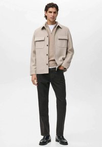 Beige wollen blend shacket met twee borstzakken, gelaagd over een lichtbeige trui. Donkere jeans en zwarte veterschoenen maken de outfit compleet.